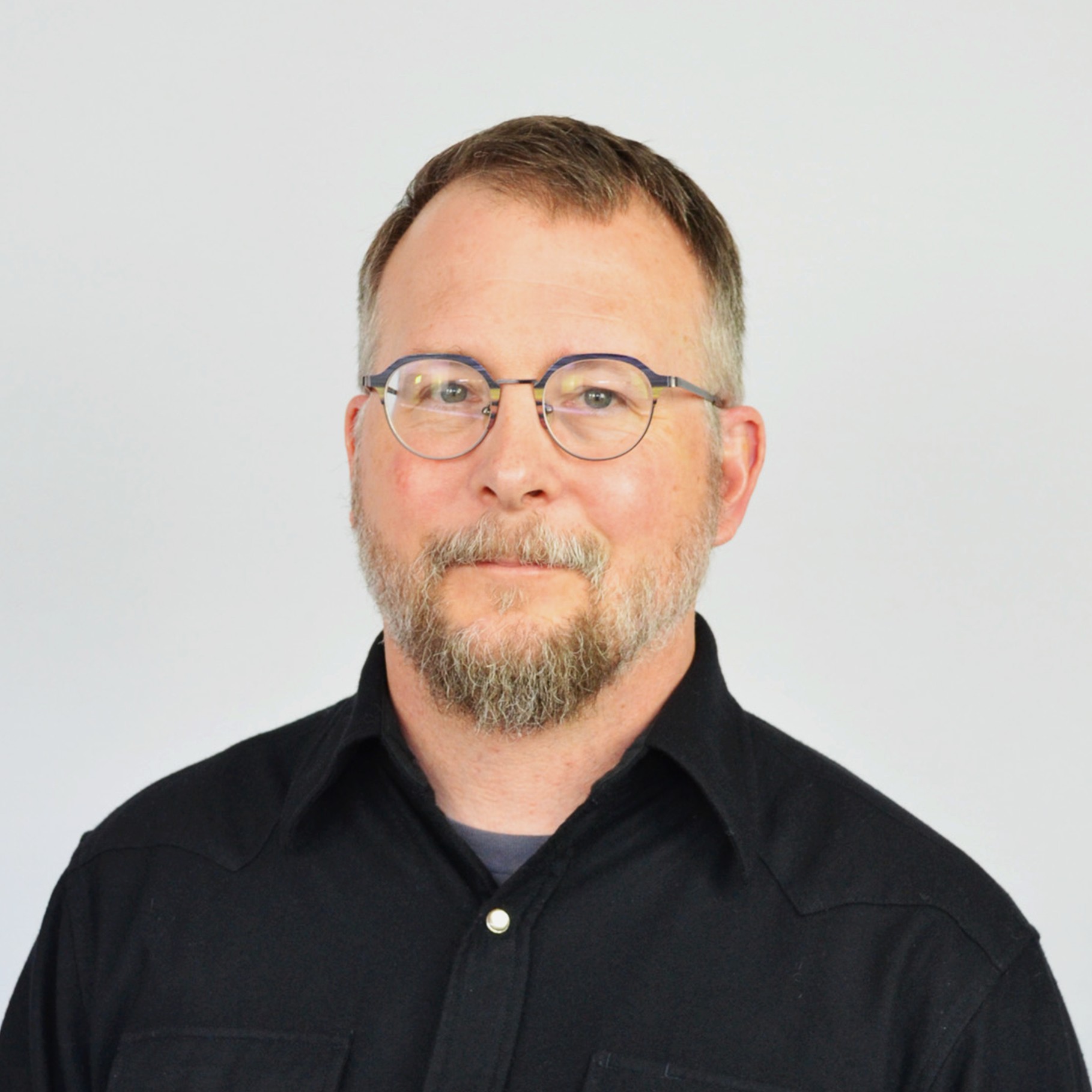 Man in Black Shirt with Short Hair and Beard on White Background Man wearing a black button-up shirt with short hair and a trimmed beard against white background.