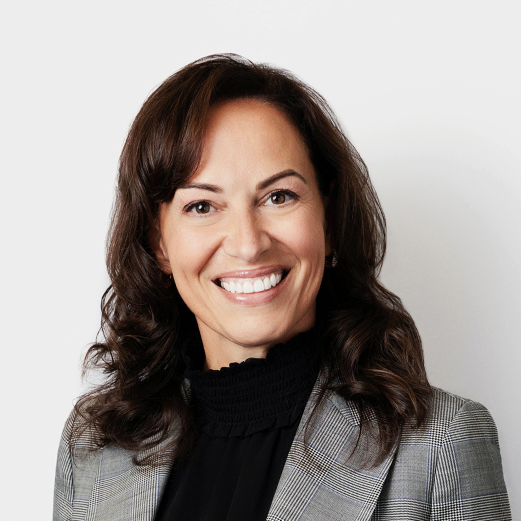 A woman with wavy brown hair wearing a gray plaid blazer and a black high-neck blouse.
