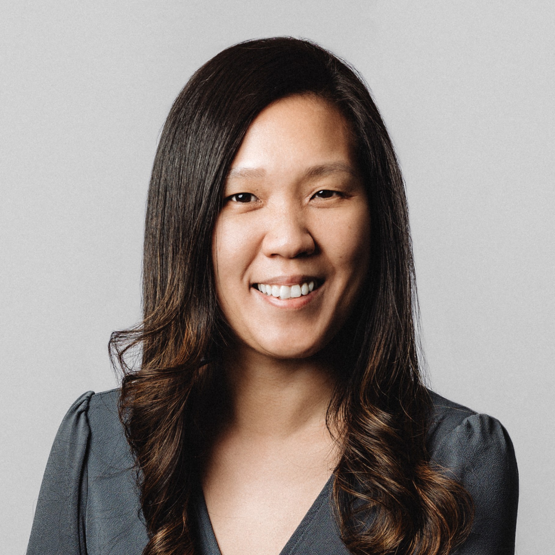 Woman with long wavy hair wearing a dark gray blouse on gray background Portrait of a woman with long dark brown hair wearing a gray blouse against a plain background.