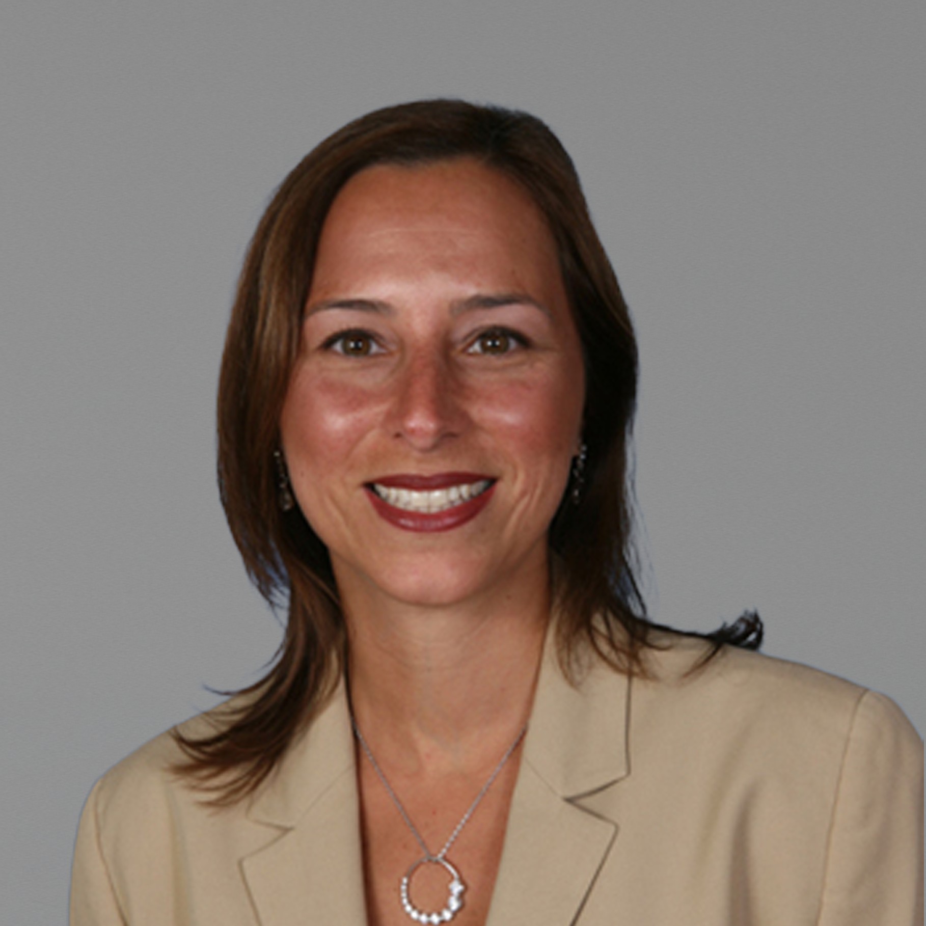 Professional woman wearing a beige blazer and a silver pendant necklace against gray.