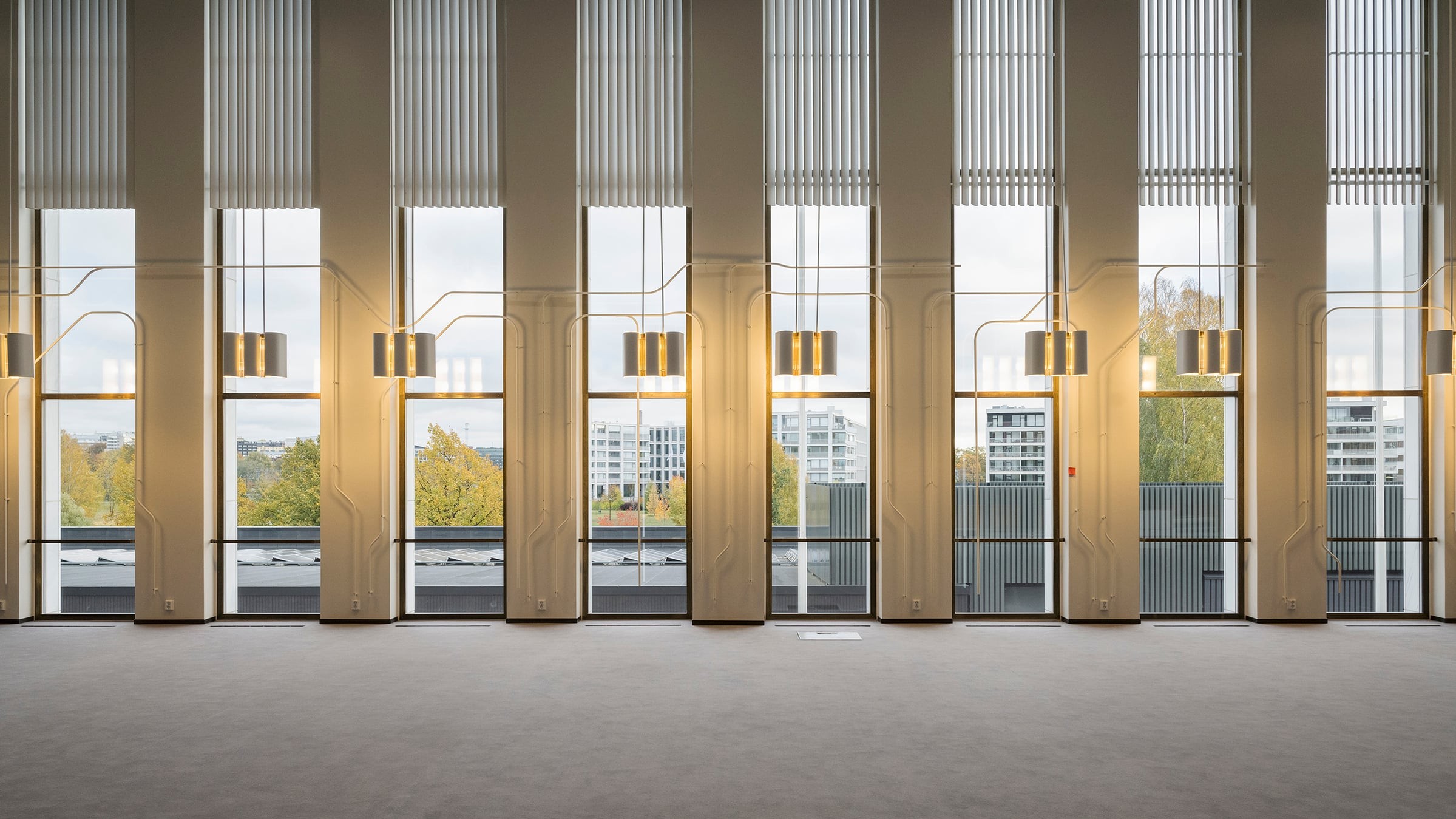 Modern interior with tall windows, vertical blinds, and pendant lights, overlooking urban greenery.