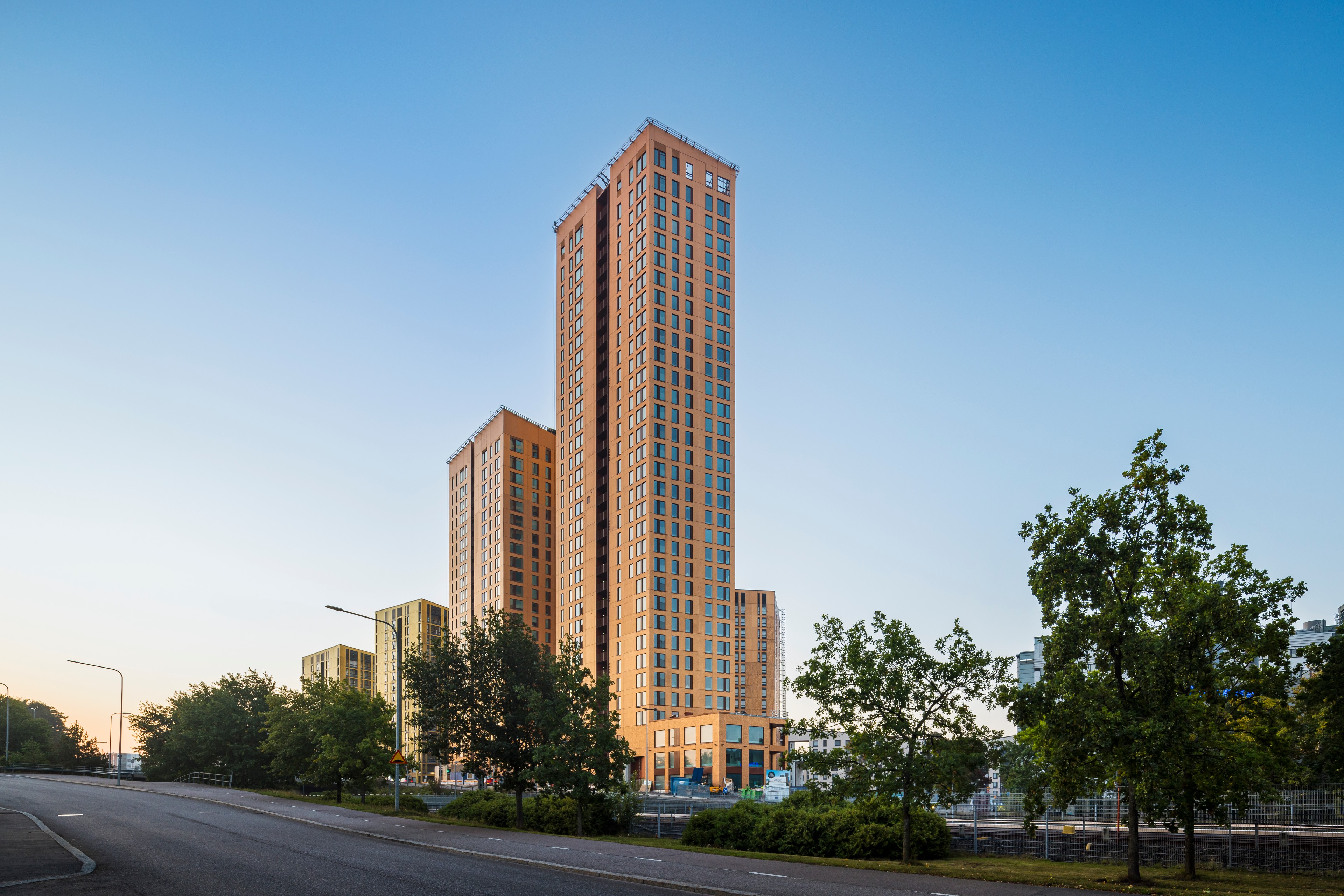 Tall modern apartment buildings with orange facades under a bright blue sky, surrounded by trees.
