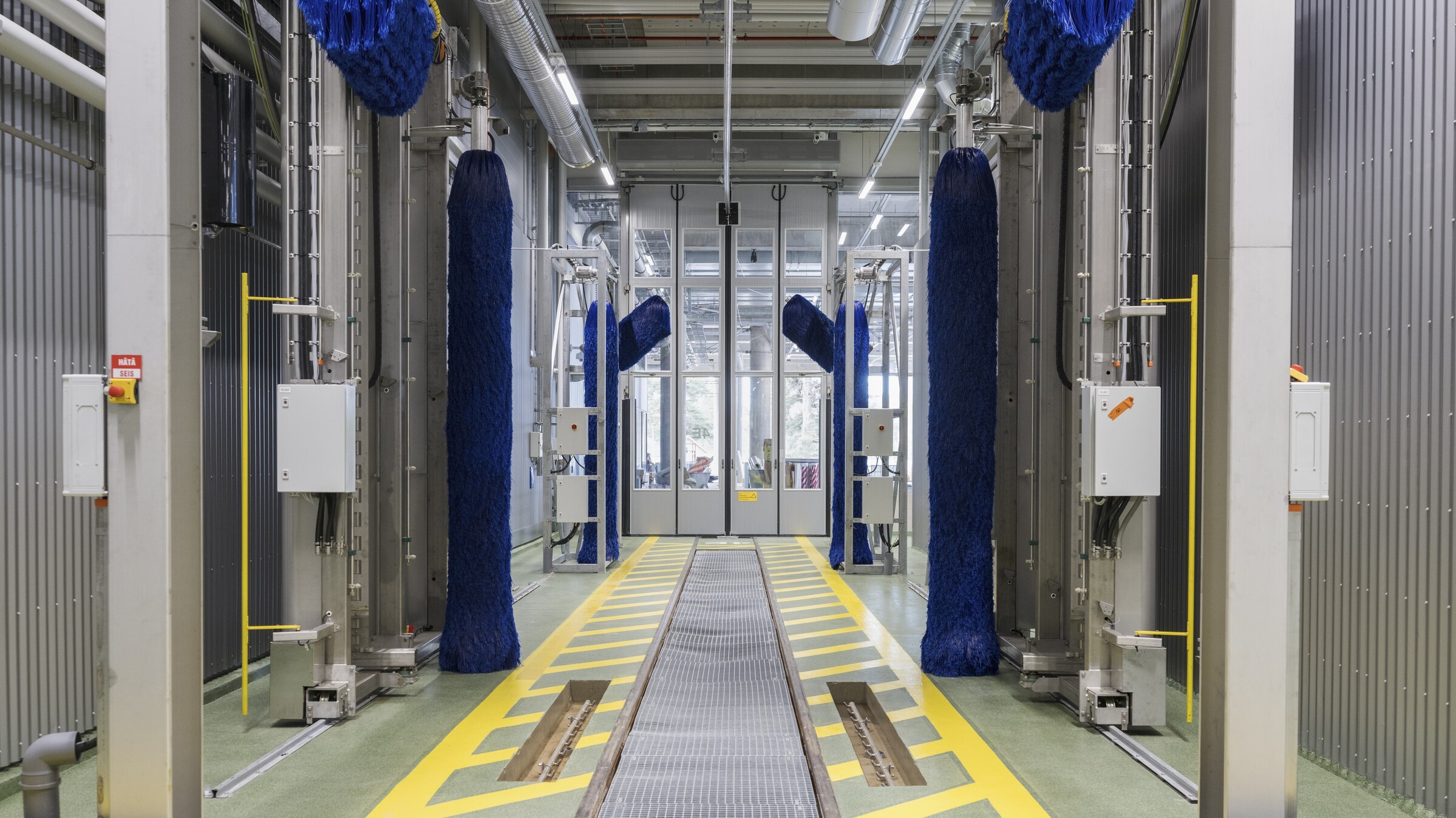 Industrial car wash facility with blue brushes, yellow safety markings, and control panels.