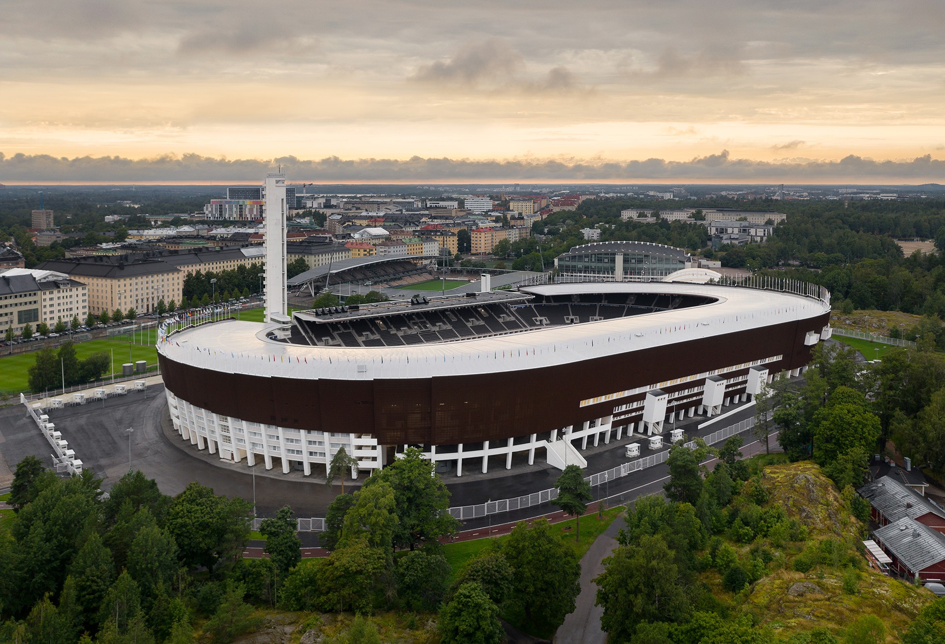 Aerial view of an oval stadium surrounded by trees, set against a cityscape under a cloudy sky.