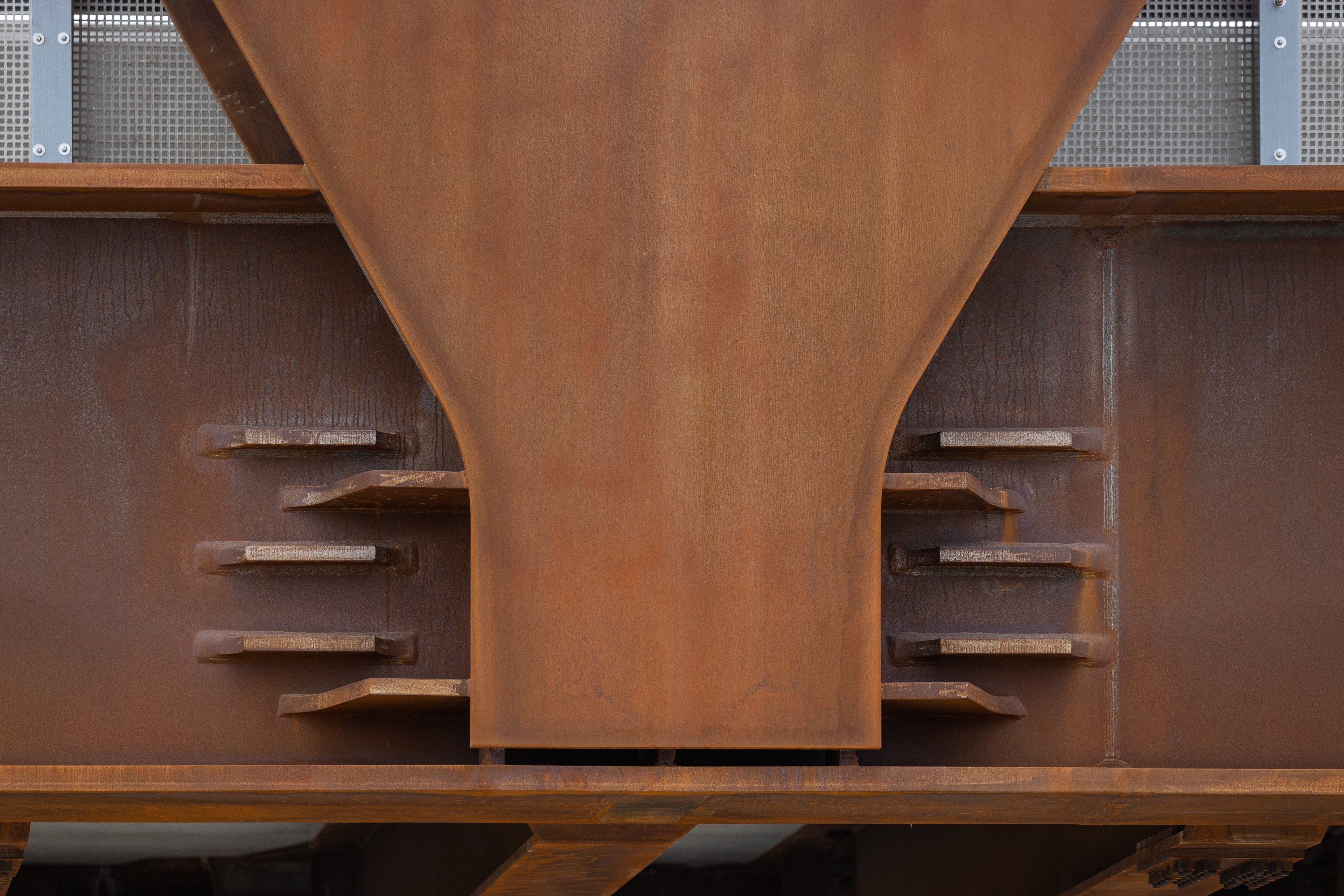 Rusty metal beam with bolted joints and a perforated panel in the background.