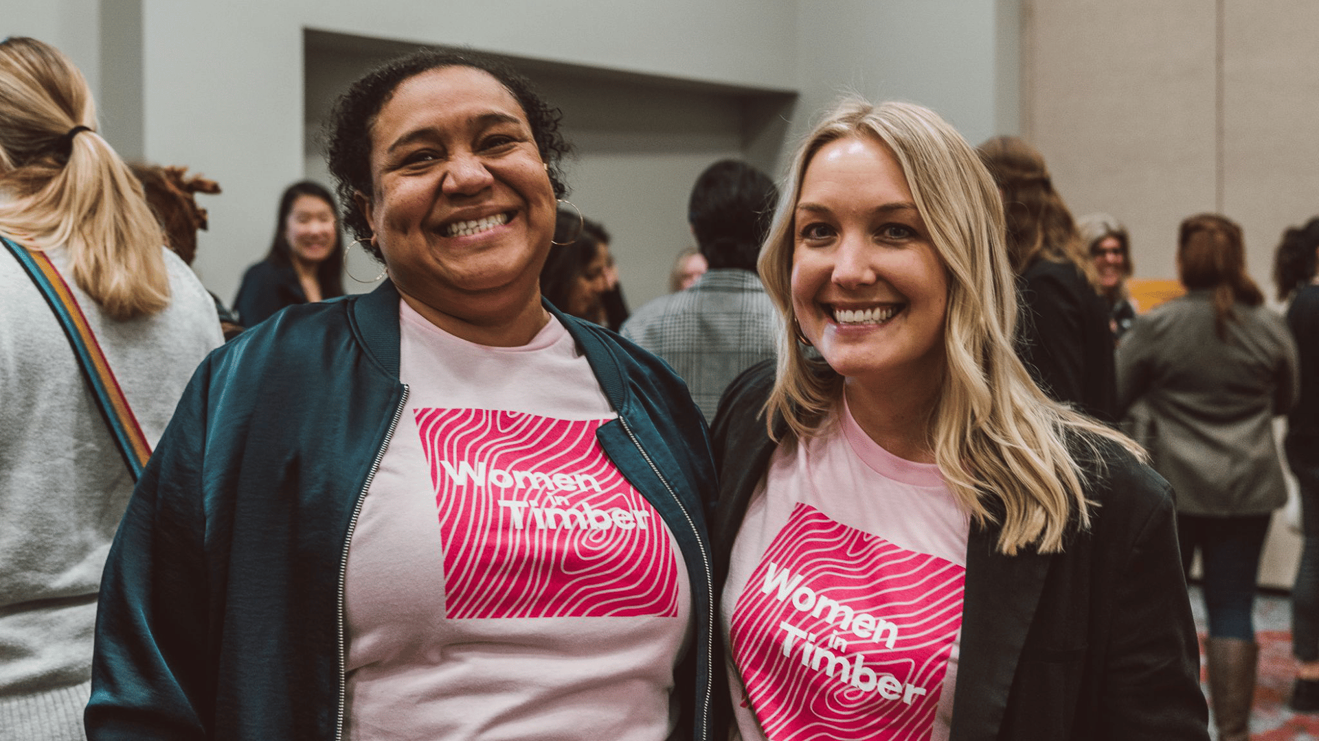 Two women wearing matching "Women in Timber" pink shirts at an indoor networking event.