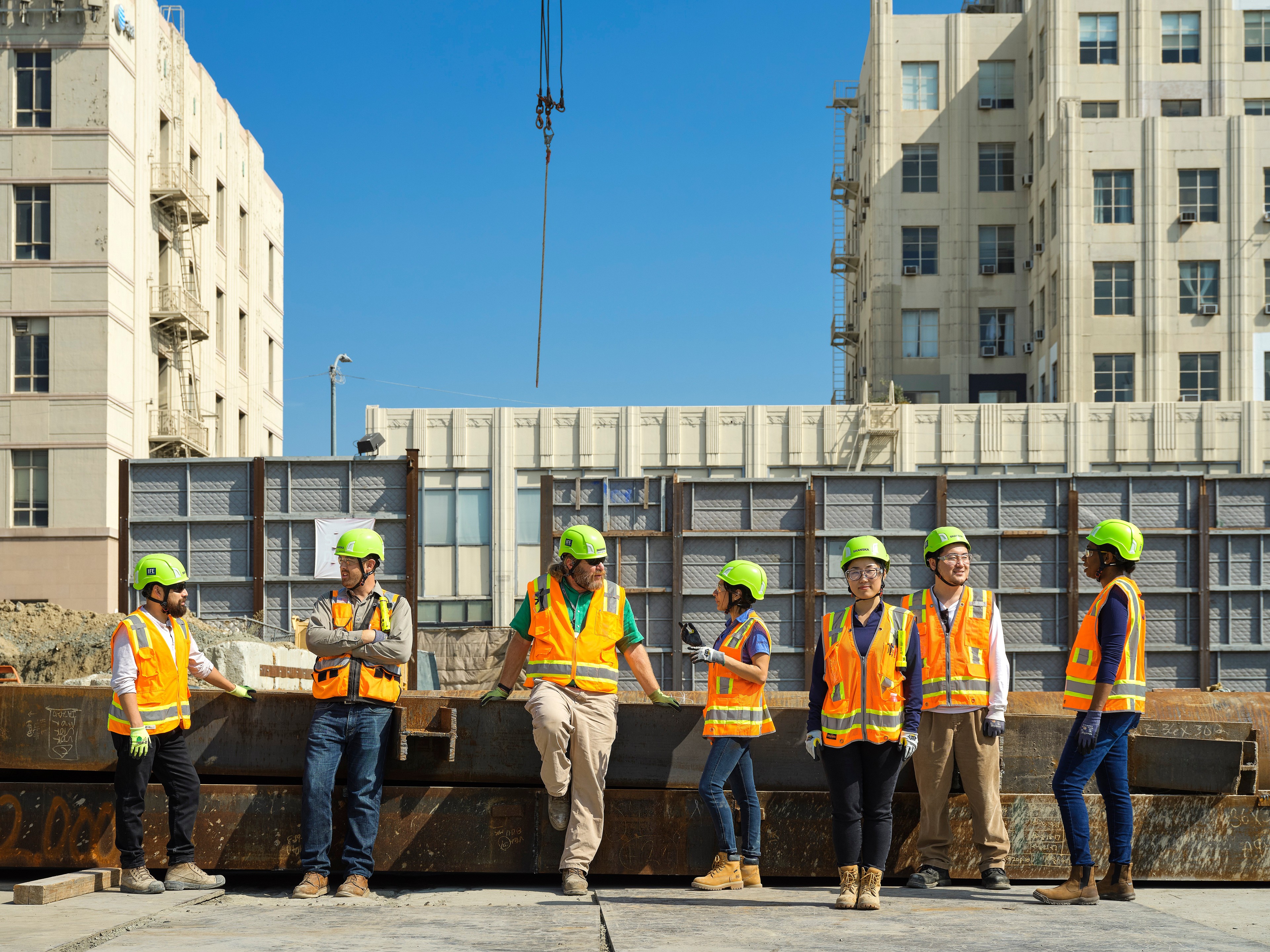 Construction workers in safety gear stand on a site with buildings in the background.