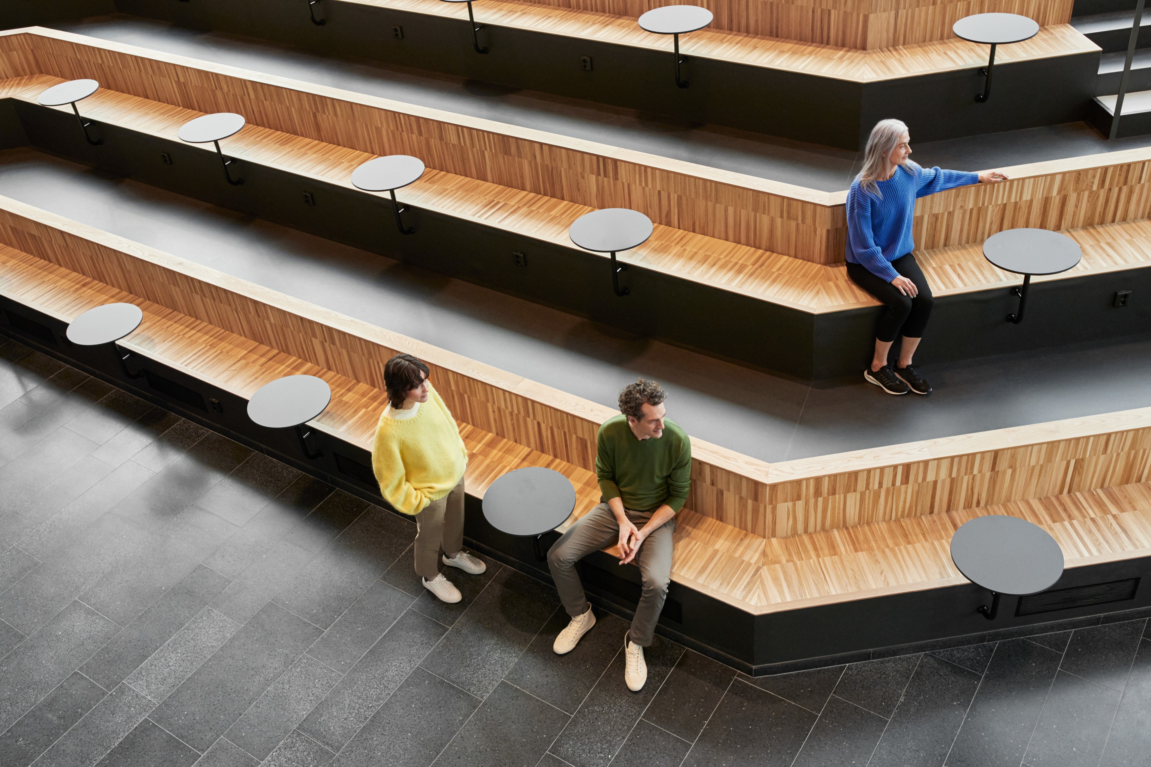 Three people in colorful sweaters sit and stand on tiered wooden seating with round tables.
