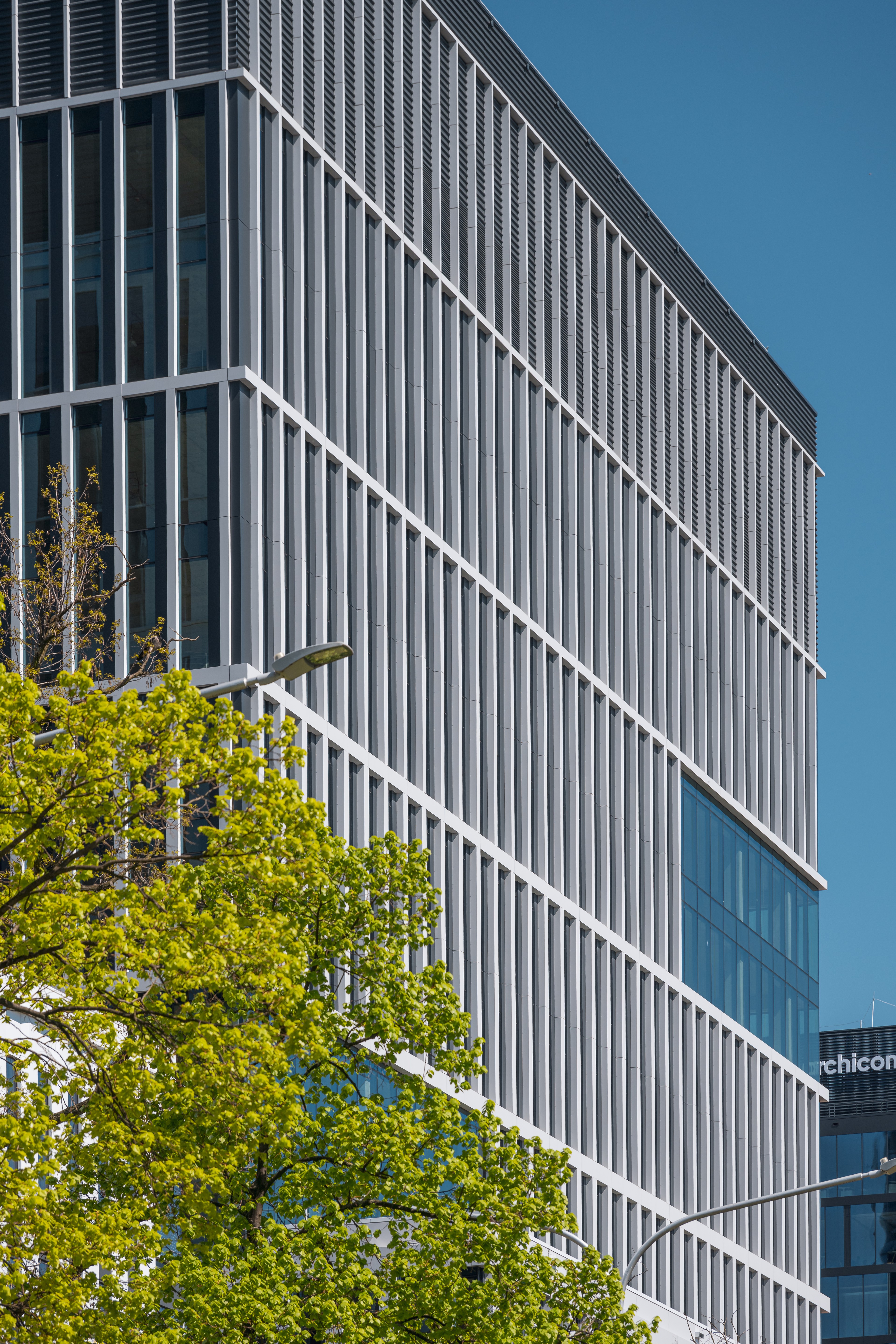 Modern office building with glass windows and green trees Modern office building with vertical white panels and a green tree in the foreground under a blue sky.