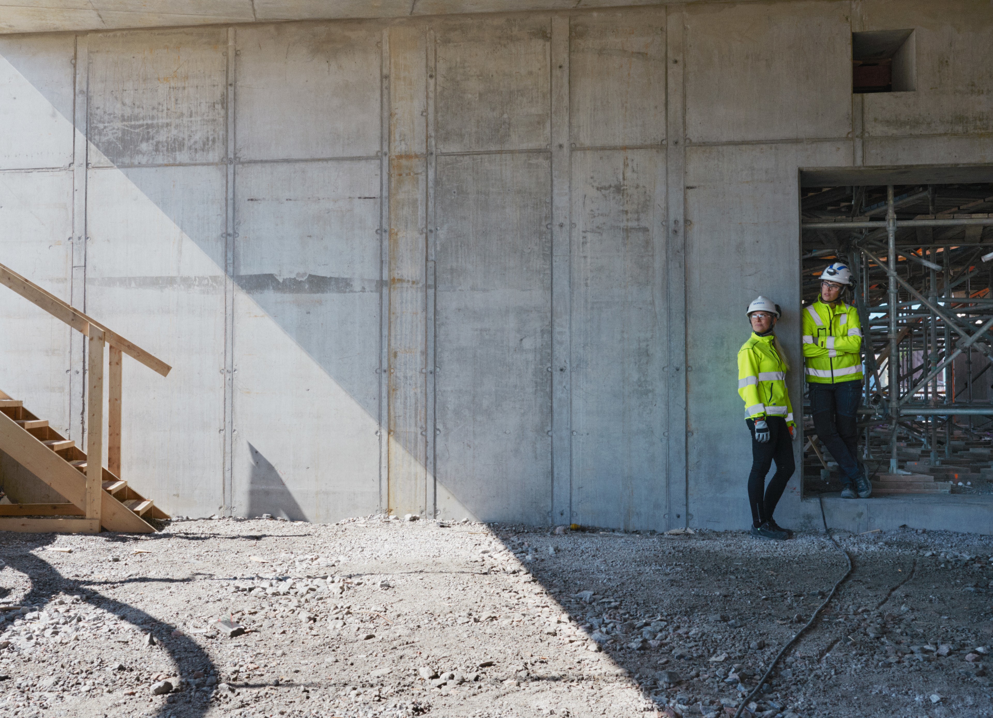 Two construction workers in safety gear stand by a concrete wall at a building site.