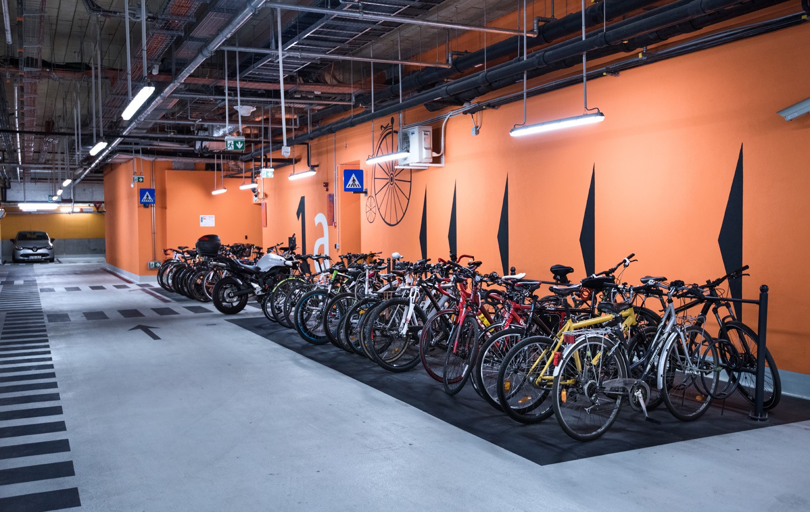 Indoor bicycle parking area with multiple bikes secured in racks against an orange wall.
