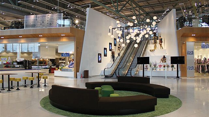 Modern shopping mall interior with escalators, seating area, and hanging lights in a spacious atrium.