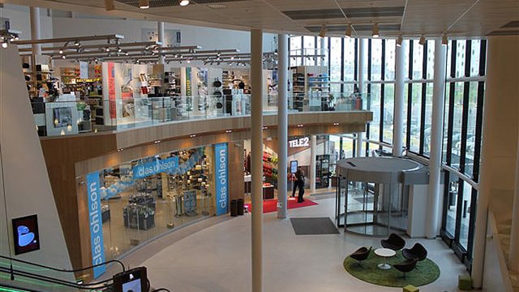 Modern shopping mall interior with glass walls, multiple stores, and seating area near entrance.