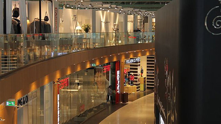 Modern shopping mall interior with glass railings and various clothing store displays.