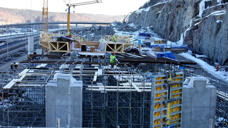 Construction site with scaffolding and cranes, building a structure near a rocky hillside in winter.