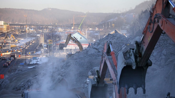 Excavators move rubble on a busy construction site, with distant hills and traffic in the background.