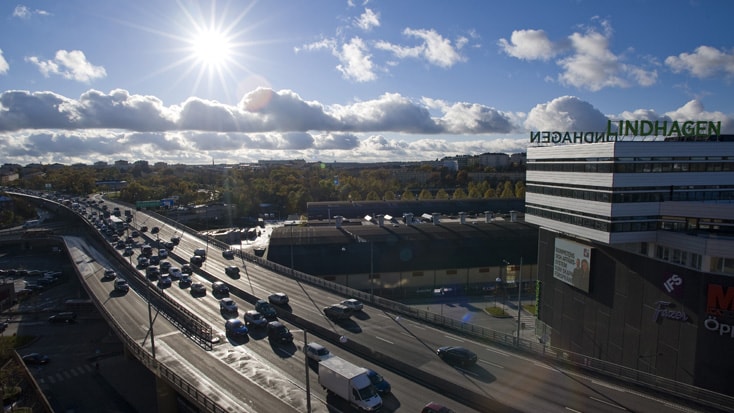 Busy highway under a bright sun with modern buildings, including one labeled "LINDHAGEN."