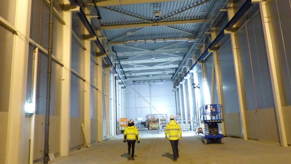 Two people in safety gear walk through a large industrial warehouse under construction.