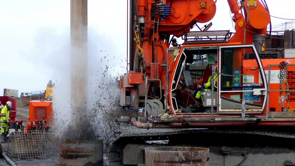 Construction site with heavy machinery drilling into the ground, releasing water and debris.