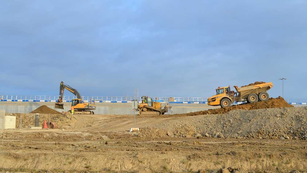 Construction site with excavators and dump trucks moving earth near a large barrier wall.