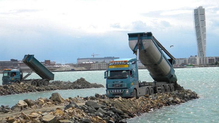Norra Hamnen Dump trucks unloading rocks into the sea, with a cityscape and skyscraper in the background.