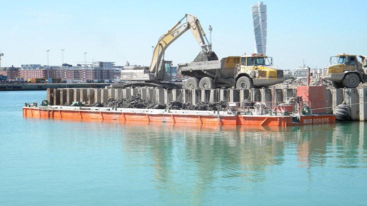 Norra Hamnen Excavators loading soil onto trucks on a barge in a harbor, with urban buildings in the background.