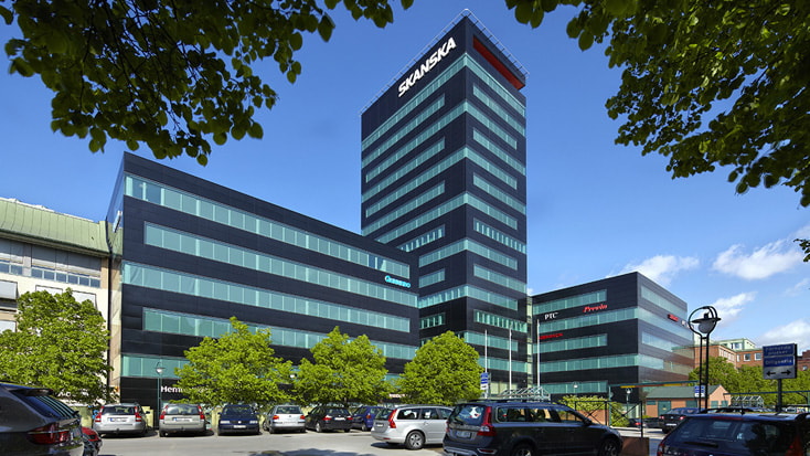 Modern office building with Skanska branding and multiple cars parked in front under a clear sky.