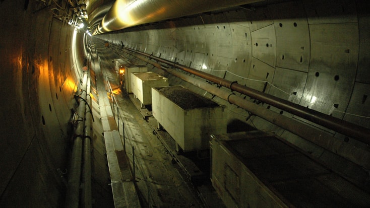 Hallandsås Construction tunnel with concrete walls, pipes, and rail carts, illuminated by overhead lighting.