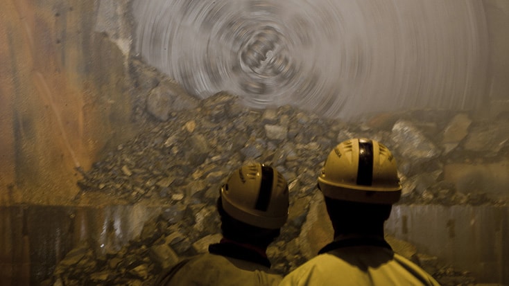 Hallandsås Two workers in helmets observe a large tunnel boring machine cutting through rock underground.