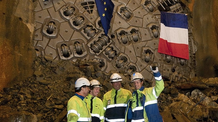 Workers stand in front of a large tunnel boring machine adorned with European and French flags.
