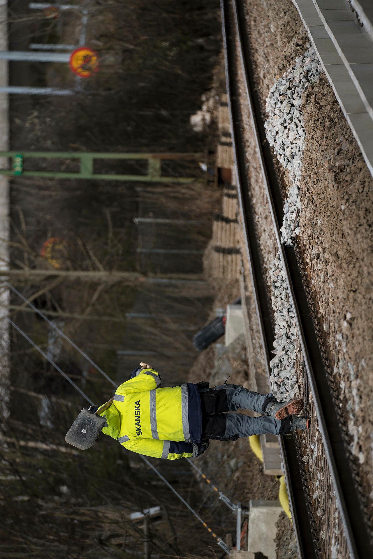 Production started in February 2020. Construction worker in a Skanska jacket walking along a railway track, carrying a shovel.