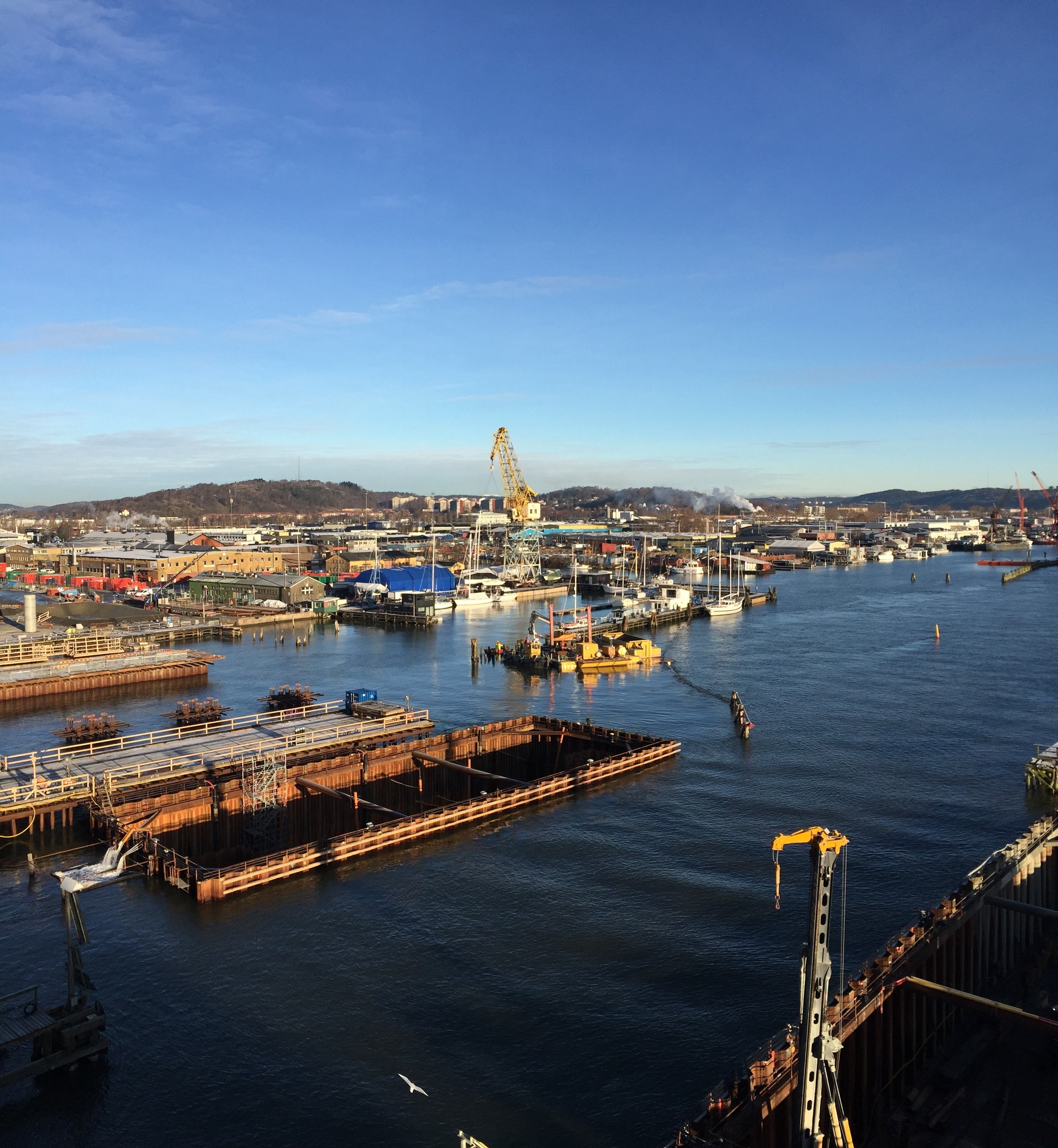 A bustling harbor scene with cranes, ships, and industrial buildings under a clear blue sky.