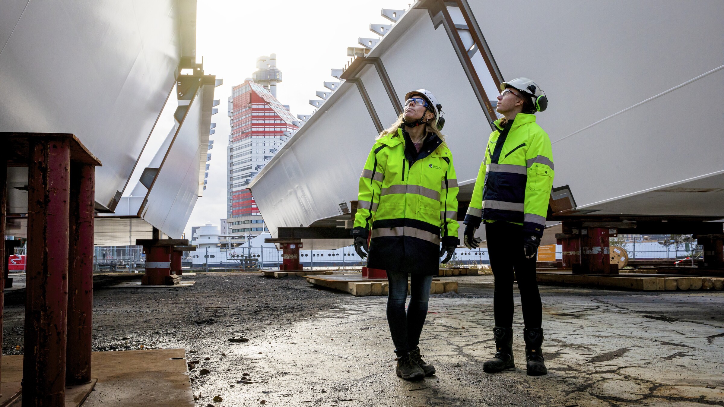 Two workers in safety gear inspect large industrial structures outdoors, with a city building in view.