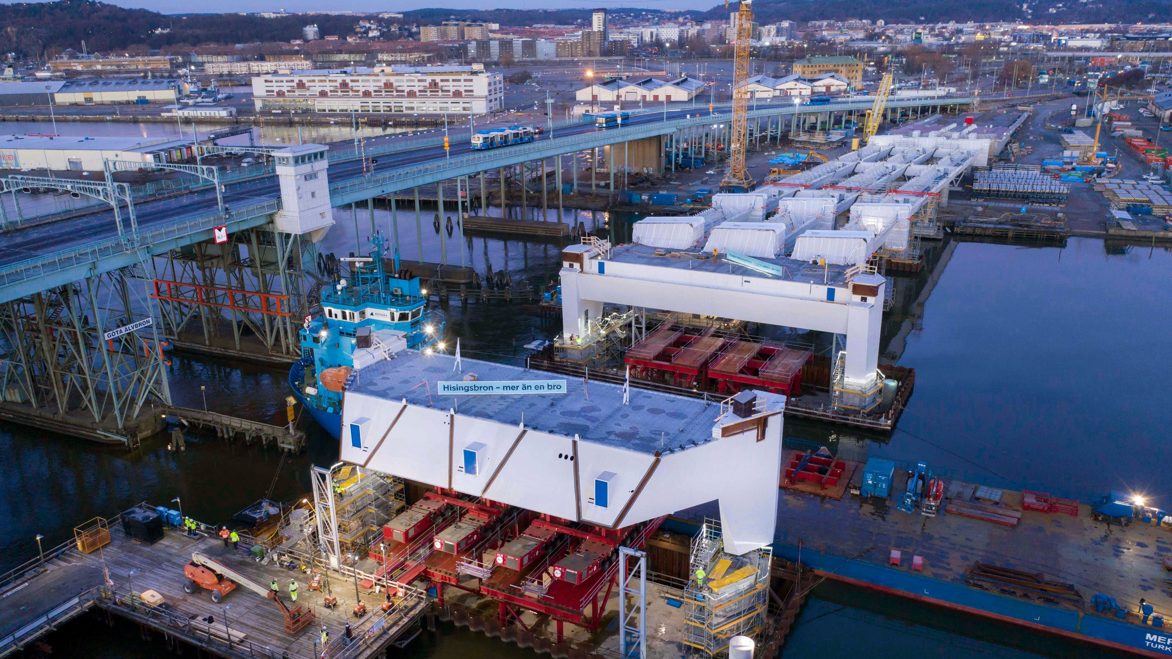 Aerial view of a bridge construction site with cranes and workers, surrounded by water and buildings.