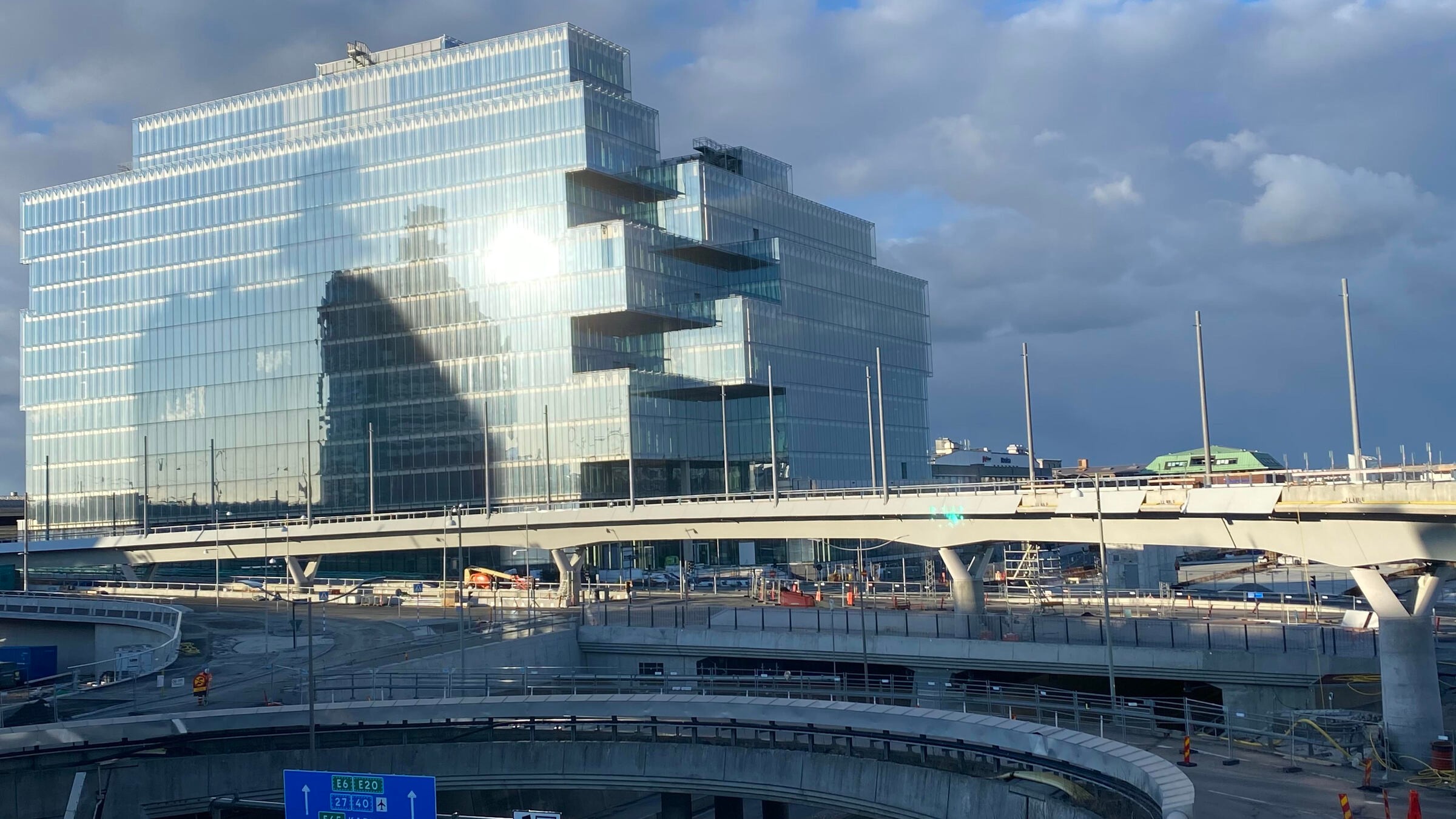 Modernes Glasgebäude mit gestuftem Design neben erhöhter Fahrbahn unter bewölktem Himmel.