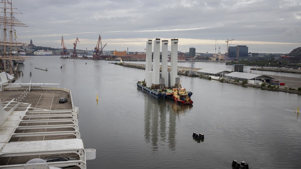 A barge carrying tall, white structures navigates a harbor with cranes and buildings in the background.