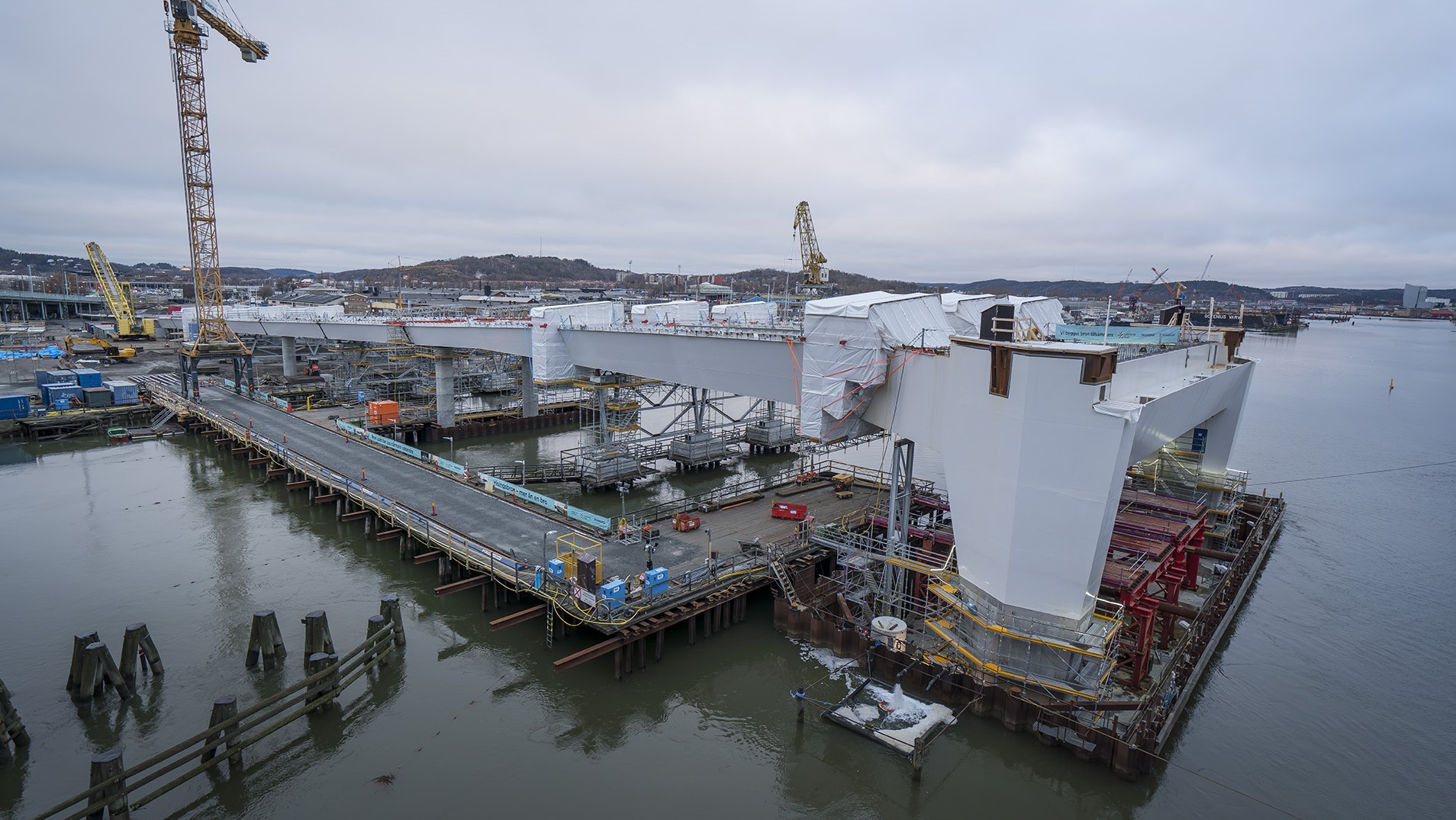 Bridge construction site over water with cranes, scaffolding, and large structural elements.