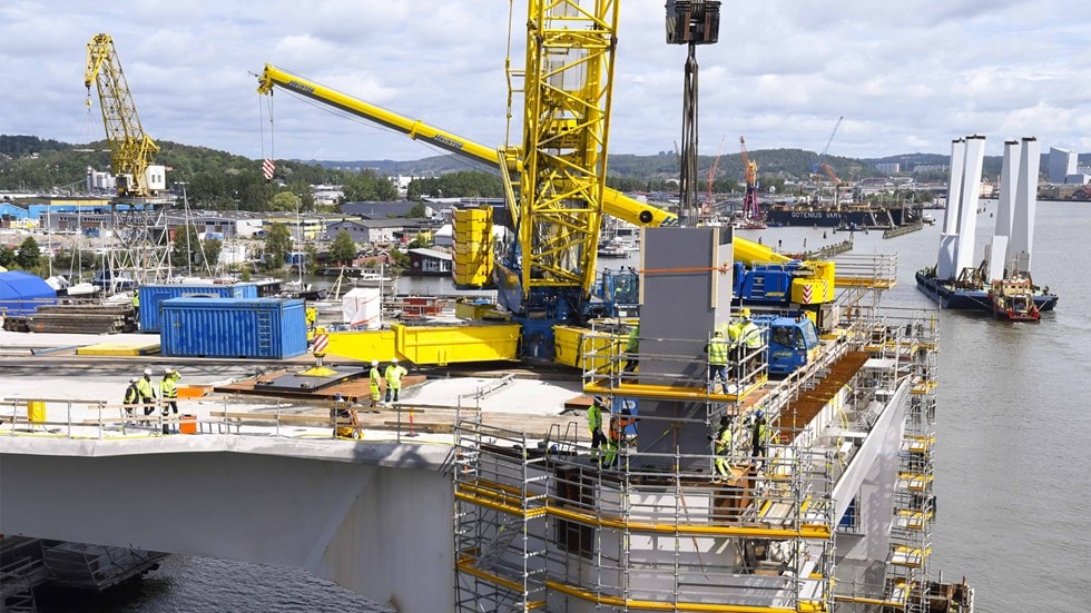 Construction workers and machinery on a bridge project by a waterfront, with cranes and scaffolding.