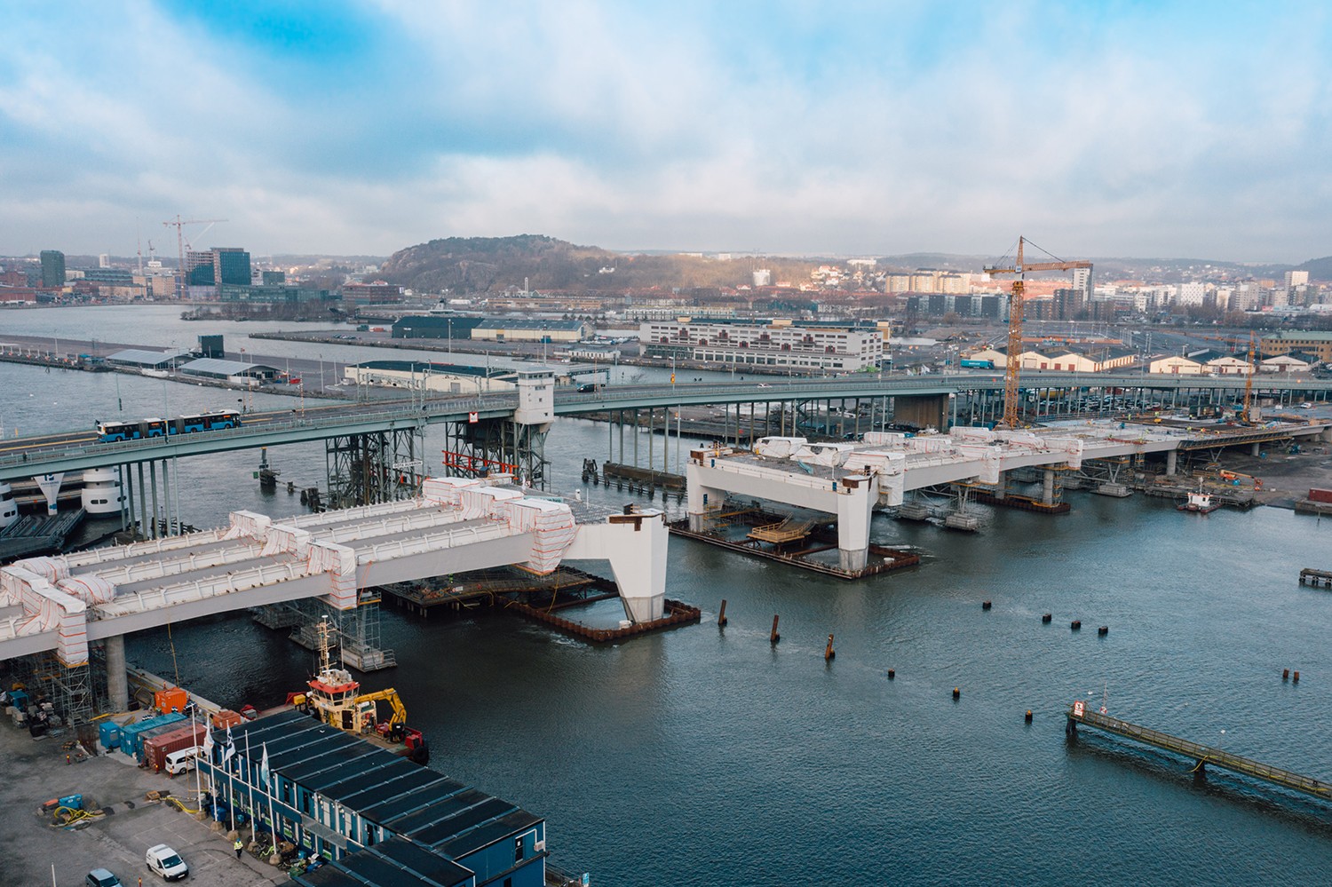 Construction of a bridge over water with cranes and buildings in the background under a cloudy sky.