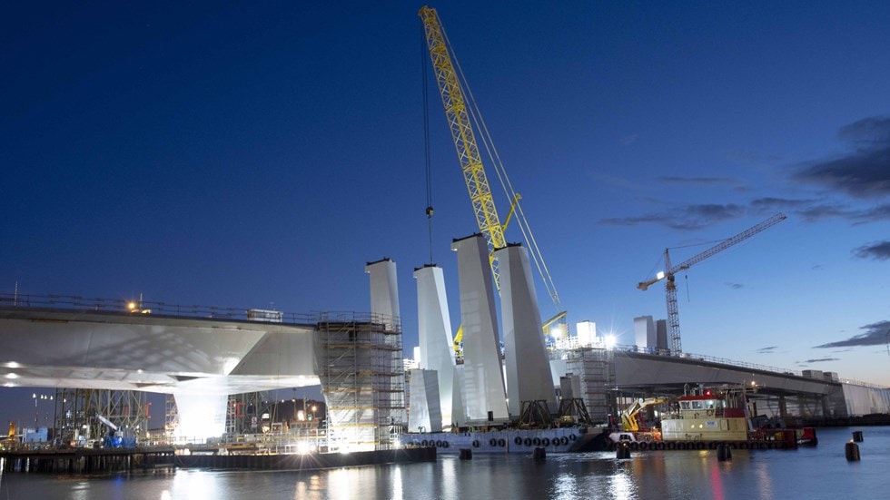 Nighttime bridge construction scene with cranes and scaffolding reflecting on the water.