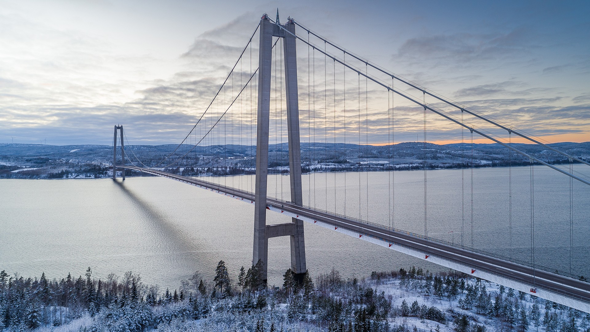 The bridge construction needs to be strong enough for the weather in northern Sweden, involving wind, ice and snow. Aerial view of a suspension bridge spanning a snowy landscape and icy river at sunset.