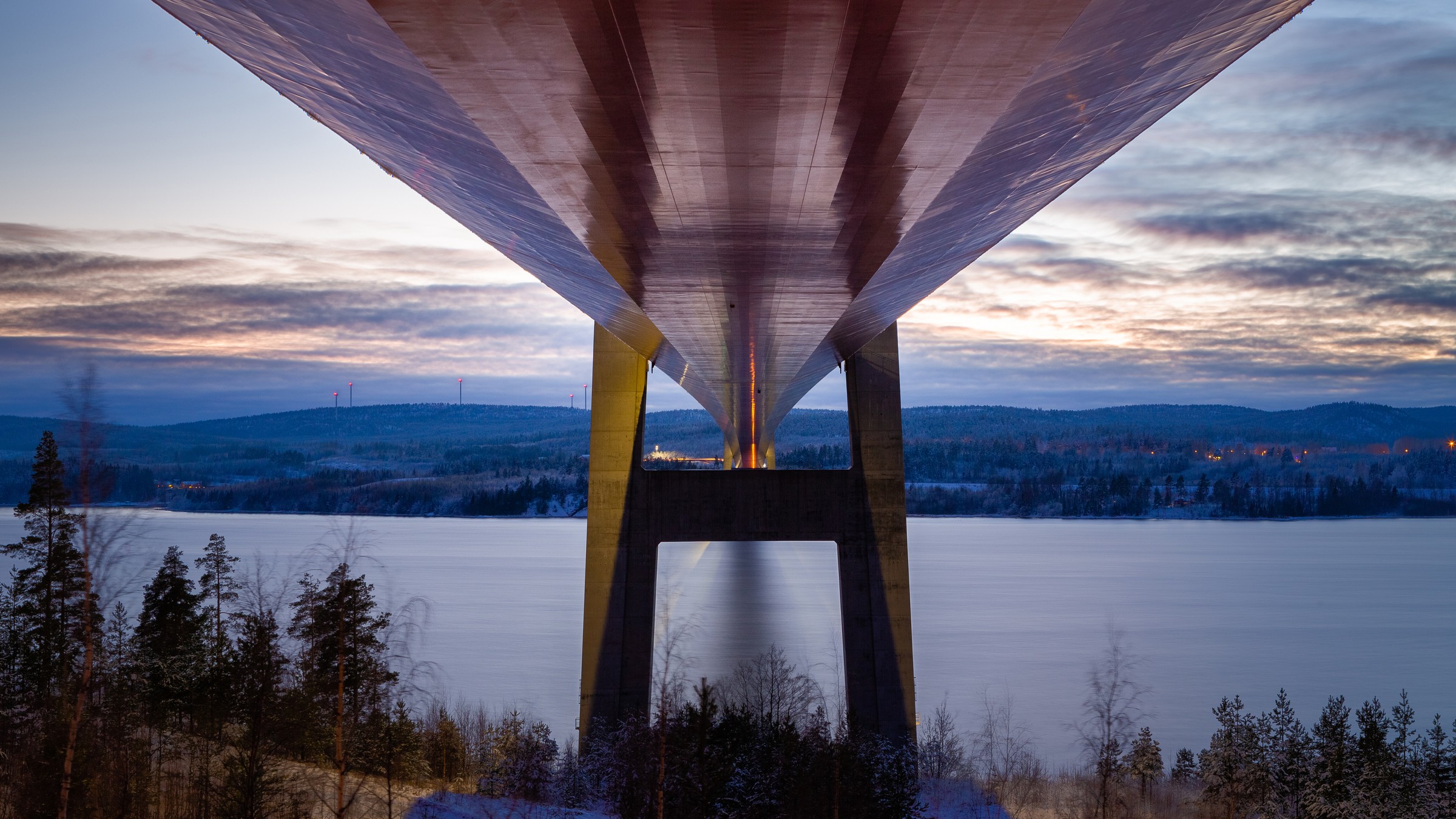 The free width of the roadway is 17.8 meters and is intended for a highway that is 13 meters wide. Underneath view of a large bridge spanning a snowy river landscape at sunset.