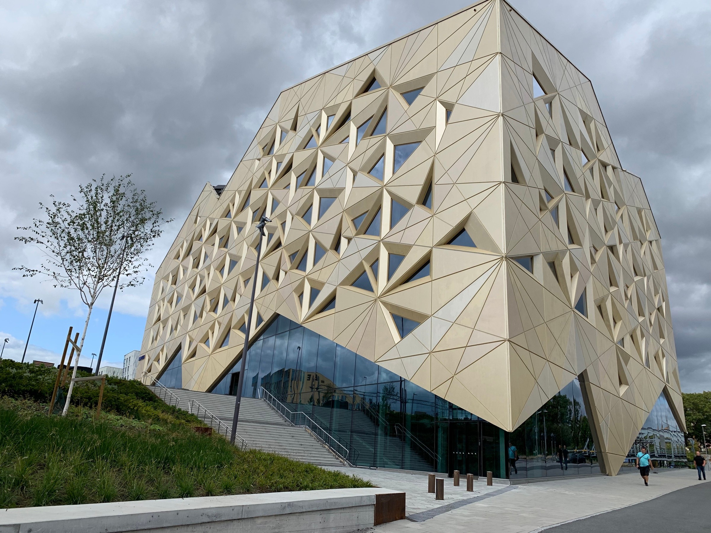 Modern geometric building with triangular patterns and glass entrance under a cloudy sky.