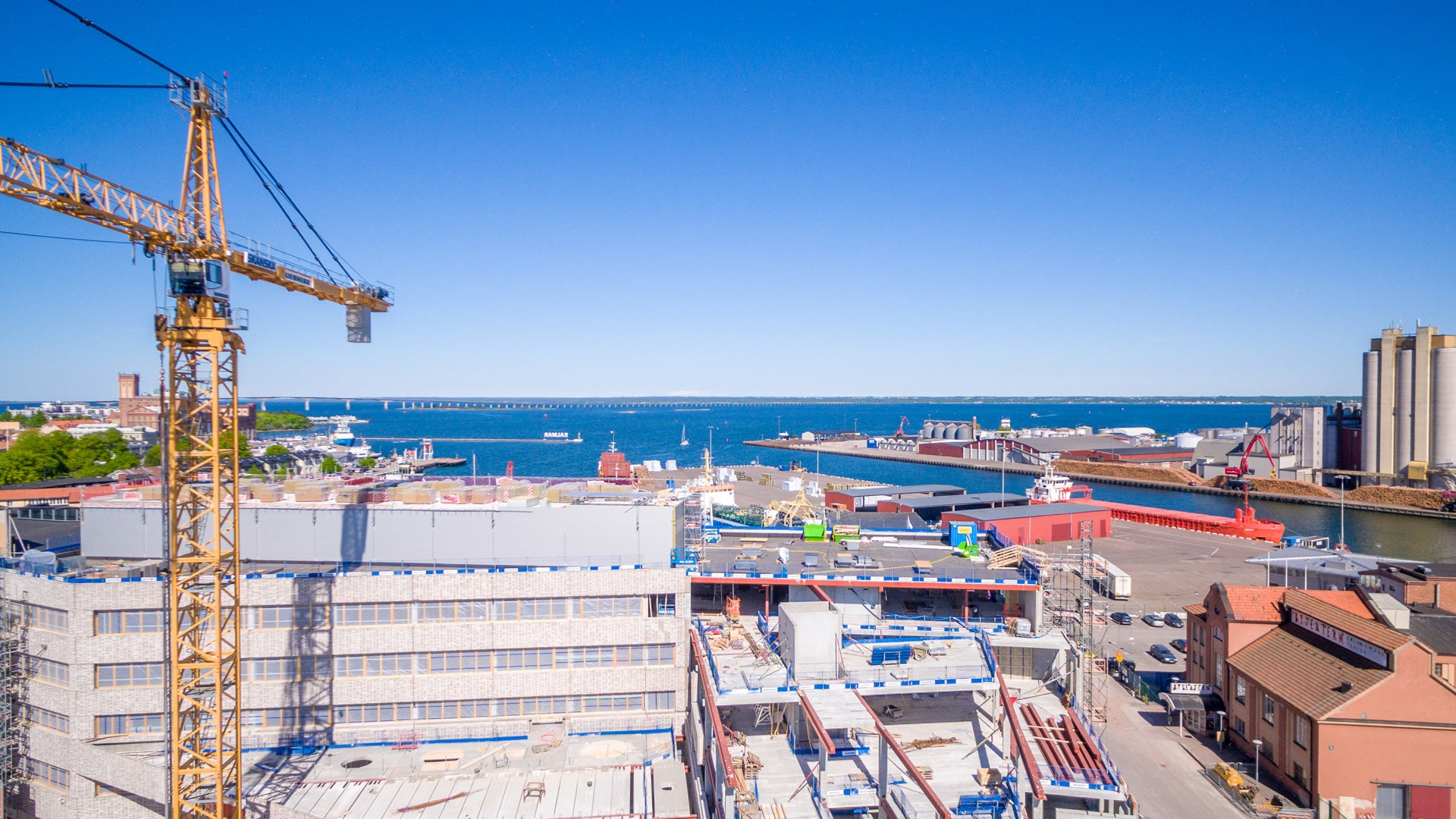 Aerial view of a coastal construction site with cranes, buildings, and a bridge over the sea.