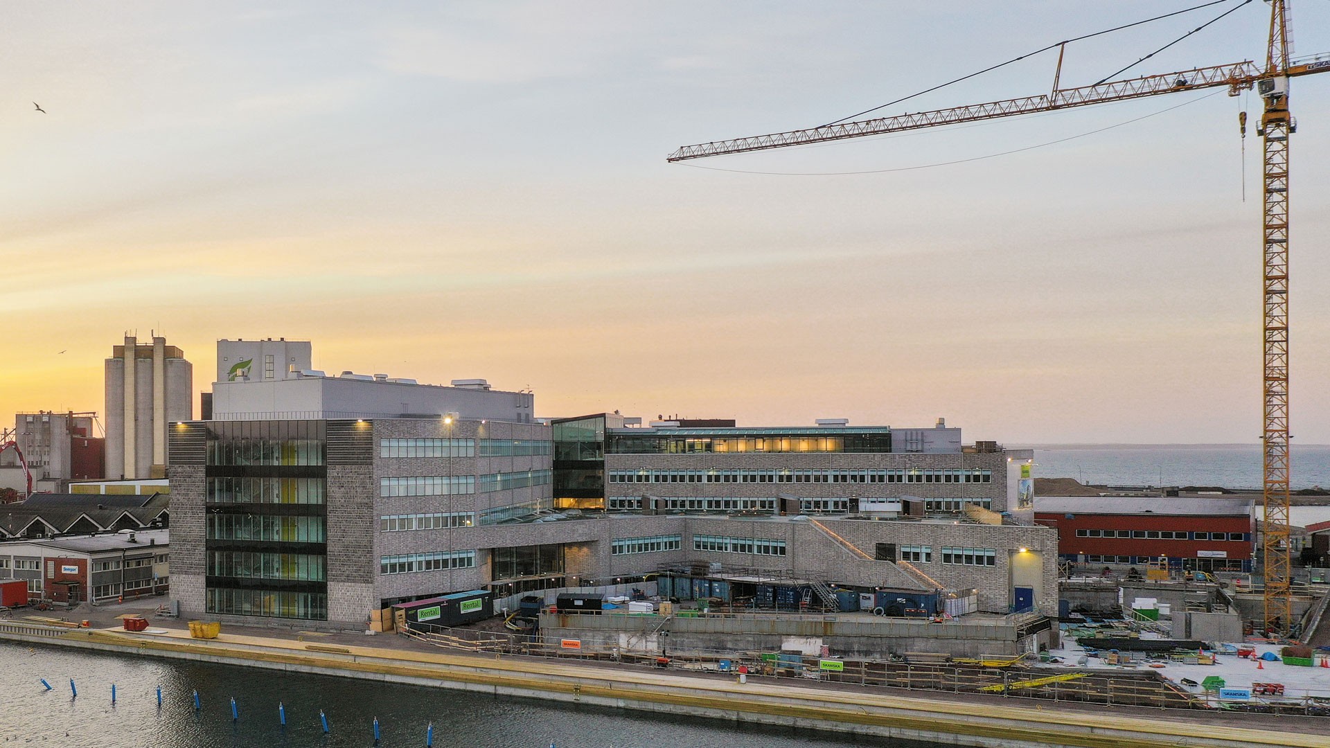 Modernes Gebäude im Bau mit einem Kran, neben einer Uferpromenade während des Sonnenuntergangs.