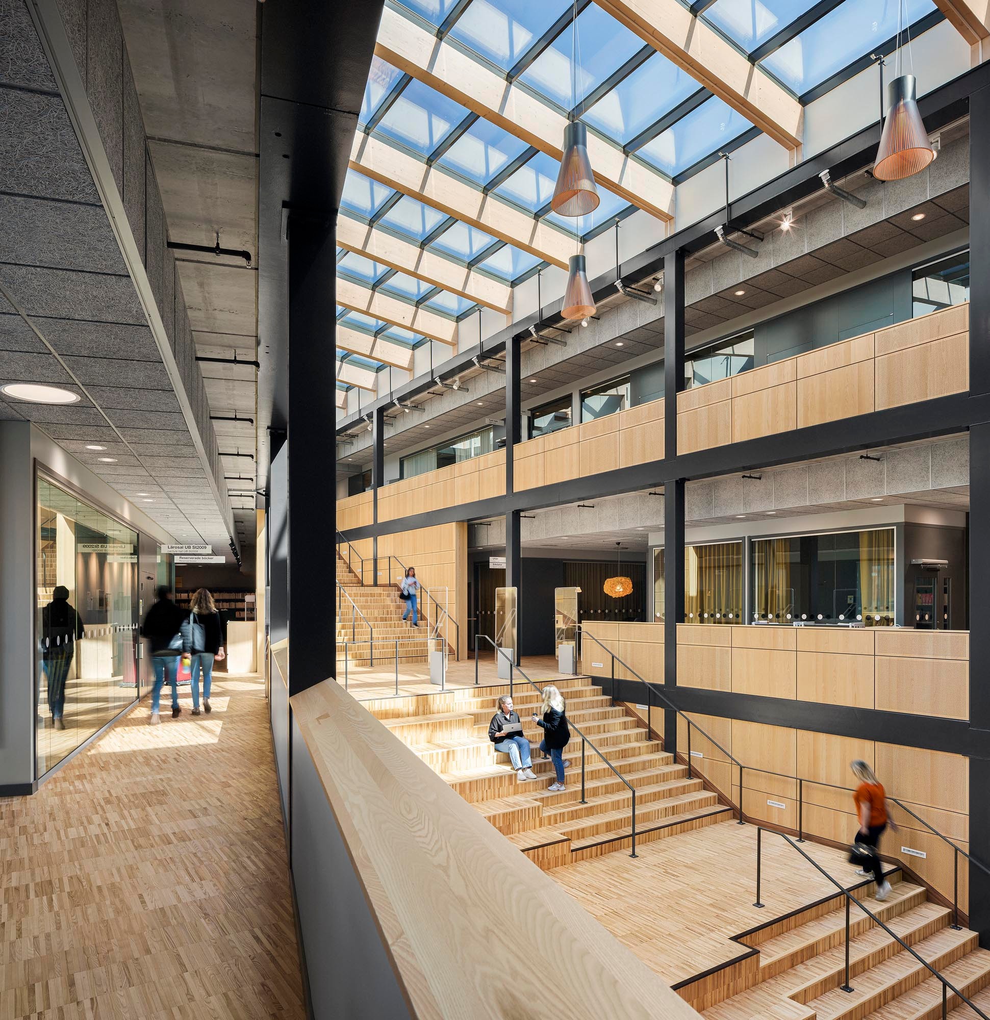 Modern atrium with wooden stairs, glass ceiling, and people walking and sitting in a spacious area.