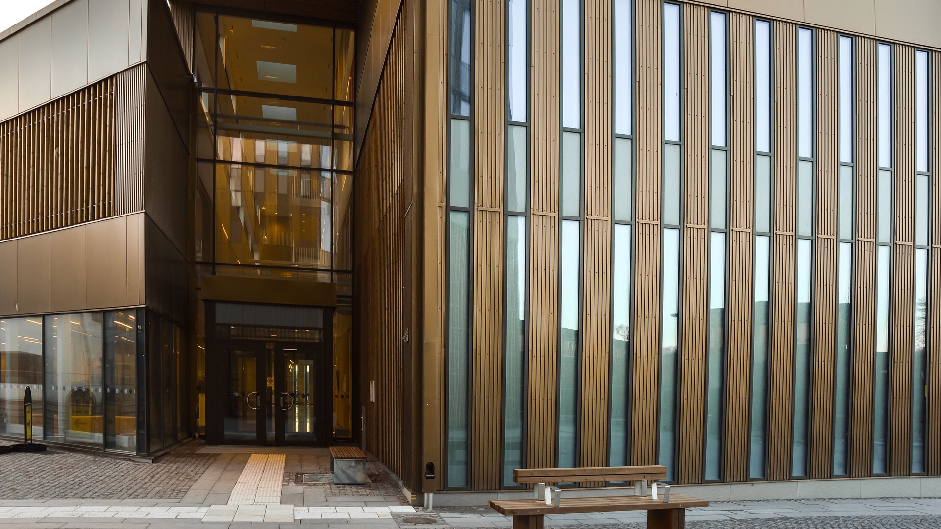 Modern building facade with tall glass and wooden panels, featuring a bench in the foreground.
