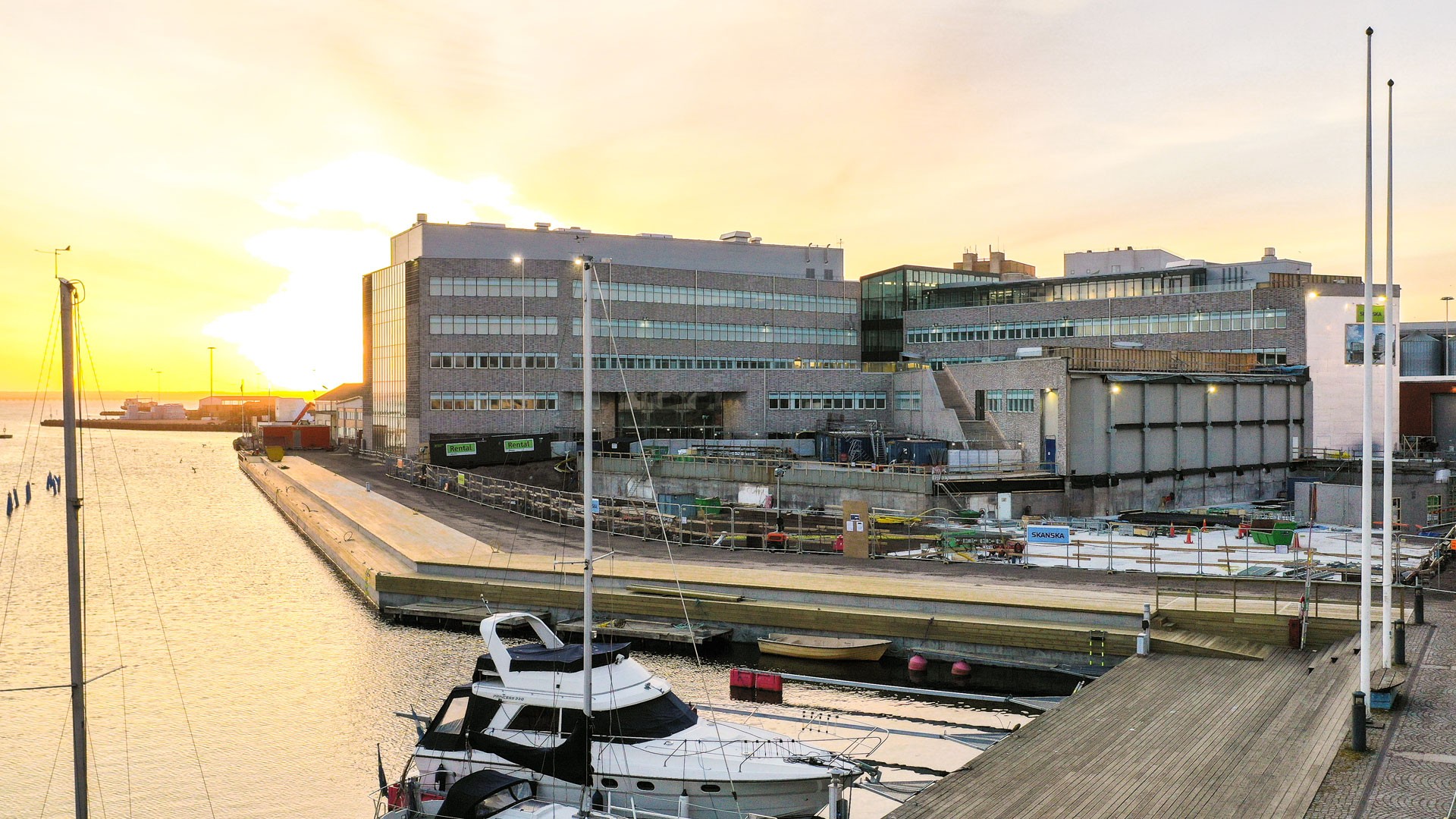 Harbor scene at sunset with docked boats and large industrial buildings in the background.