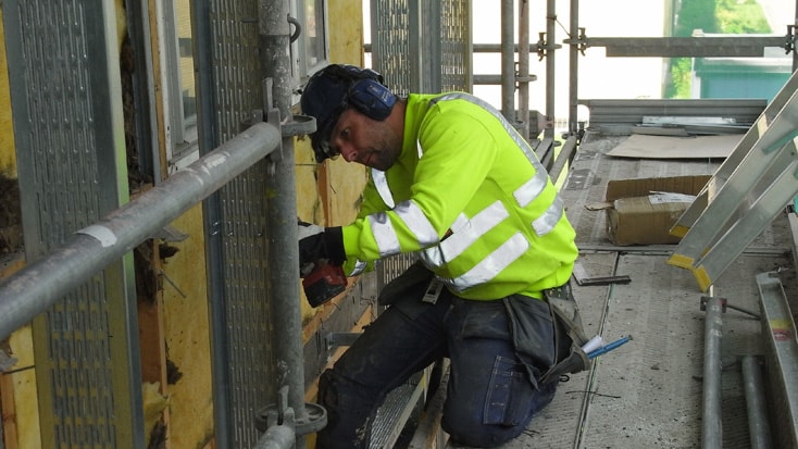 A construction worker in high-visibility gear using a drill on scaffolding at a building site.