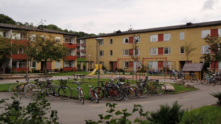 Brogården Apartment complex courtyard with bicycles, playground equipment, trees, and three-story buildings.